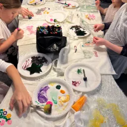 Dzieci przy stole dekorują torby bawełniane farbami i pisakami, używając szablonów i gąbek, widoczne farby na papierowych talerzach, koraliki, brokat i zabezpieczenie stołu folią.