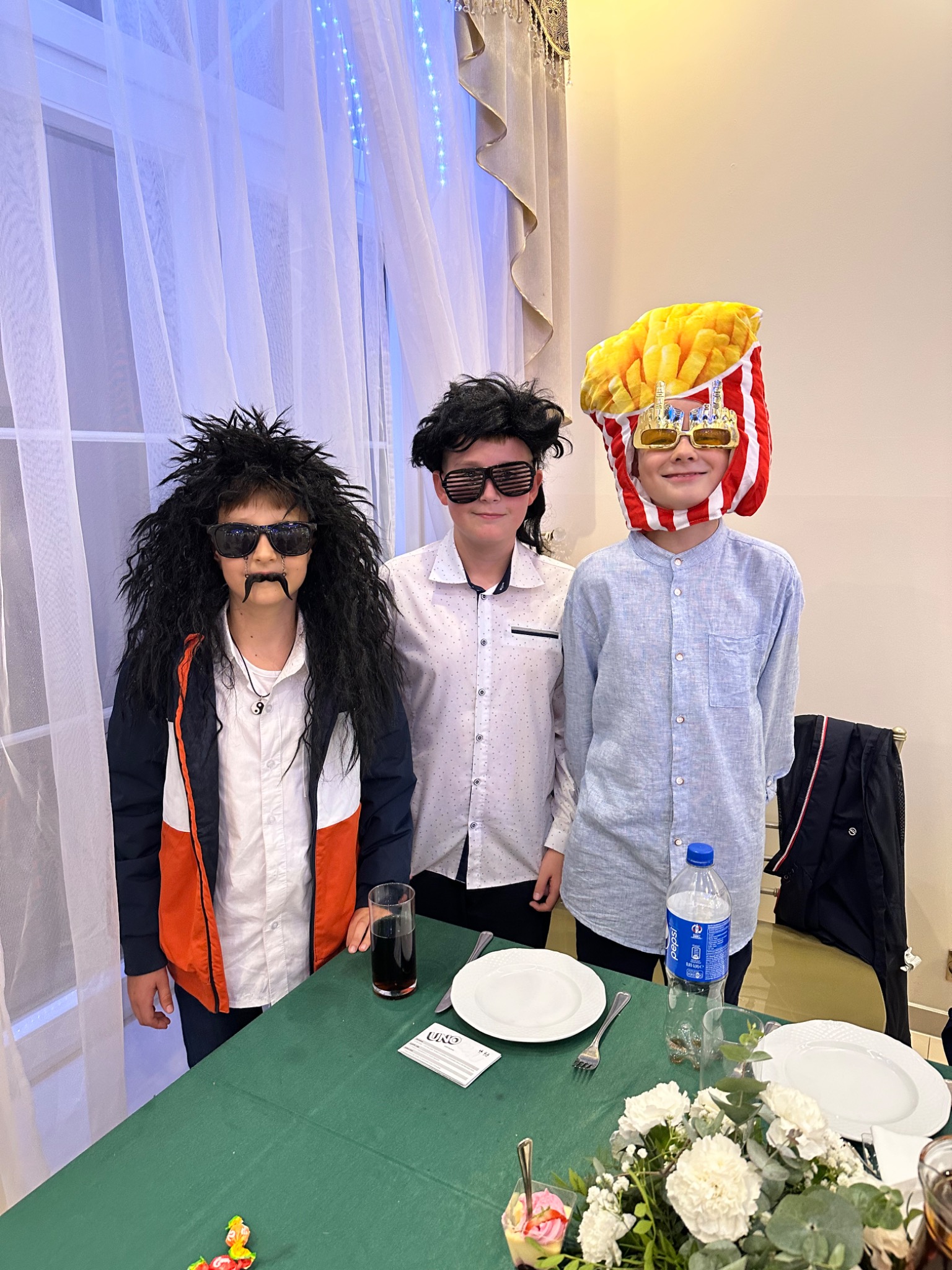 Trójka dzieci w przebraniach: jedno w peruce i doczepionych wąsach, drugie w okularach z żaluzjami, trzecie w nakryciu głowy imitującym frytki, pozują przy stole z kwiatami i napojami.