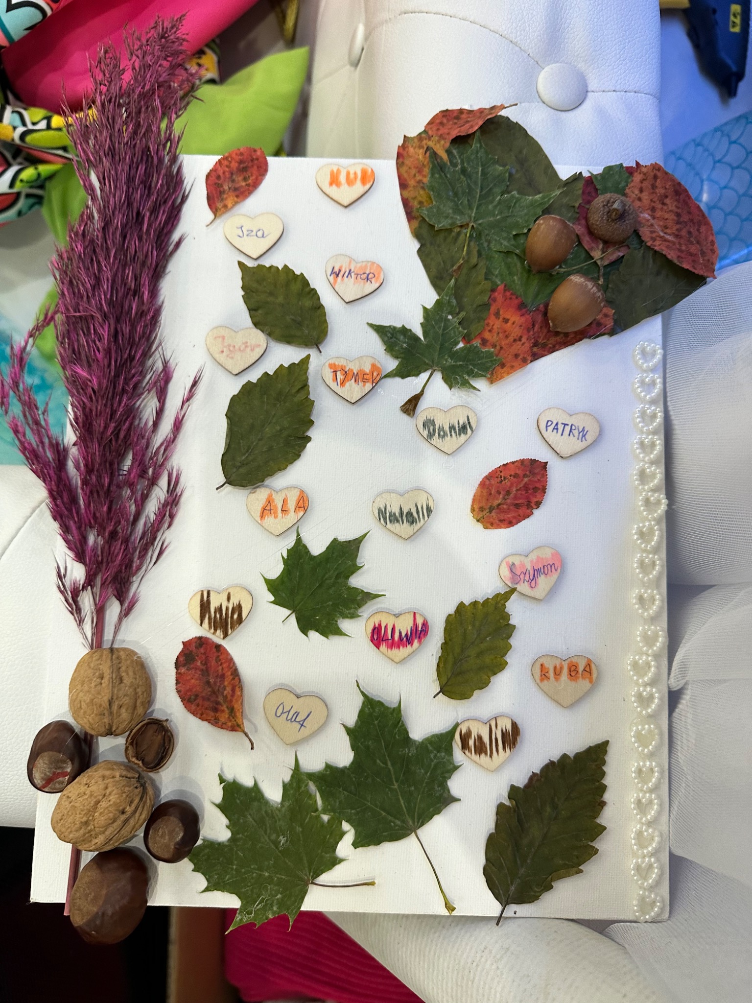 Ręcznie wykonana dekoracja z liści, kasztanów i orzechów na białym płótnie, ozdobiona drewnianymi serduszkami z imionami dzieci i perłową obwódką.