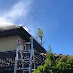 Kajetan Smoliński Nordlys - Pracownik w jaskrawej kamizelce, stojący na dachu domu z brązową dachówką, myje go myjką ciśnieniową, tworząc chmurę pary wodnej na tle błękitnego nieba. Metalowa drabina oparta o rynnę.