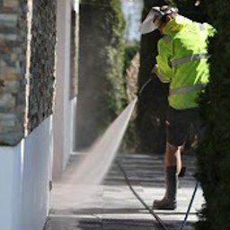 Kajetan Smoliński Nordlys - Pracownik w odblaskowej kurtce i masce ochronnej czyści kostkę brukową przy pomocy myjki ciśnieniowej, widoczny strumień wody i mokra powierzchnia kostki, elewacja budynku z elementami kamiennymi...
