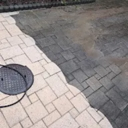 Częściowo wyczyszczona kostka brukowa z widocznym włazem kanalizacyjnym, ukazująca różnicę przed i po myciu, z zielenią w tle.