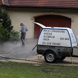 Kajetan Smoliński Nordlys - Mężczyzna w szarej bluzie czyści podjazd z kostki brukowej za pomocą myjki ciśnieniowej, obok przyczepka z reklamą usług mycia ciśnieniowego kostki, elewacji i dachów, w tle dom z czerwoną dachówką.