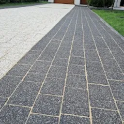 Długa, dwukolorowa ścieżka z kostki brukowej prowadząca do domu z brązową bramą garażową, z widocznym fragmentem trawnika po prawej stronie.