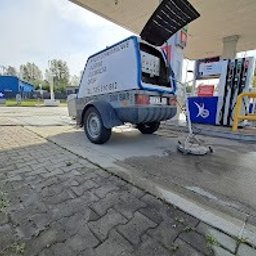 Kajetan Smoliński Nordlys - Przyczepa z urządzeniem do czyszczenia kostki brukowej na stacji benzynowej, widoczna dysza czyszcząca na kostce, błękitne logo firmy na przyczepie.