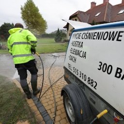 Kajetan Smoliński Nordlys - Pracownik w jaskrawożółtej kurtce i gumowcach czyści podjazd z kostki brukowej za pomocą myjki ciśnieniowej; widoczna przyczepka z napisem 'Mycie ciśnieniowe - kostka, elewacja, dach' i numerem...