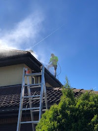 Pracownik w jaskrawej kamizelce, stojący na dachu domu z brązową dachówką, myje go myjką ciśnieniową, tworząc chmurę pary wodnej na tle błękitnego nieba. Metalowa drabina oparta o rynnę.