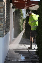 Pracownik w odblaskowej kurtce i masce ochronnej czyści kostkę brukową przy pomocy myjki ciśnieniowej, widoczny strumień wody i mokra powierzchnia kostki, elewacja budynku z elementami kamiennymi...