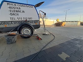 Przyczepka z napisem 'Mycie ciśnieniowe - kostka, elewacja, dach' i numerem telefonu, stojąca na wyczyszczonej części kostki brukowej, obok wiadro i narzędzia, w tle budynek.