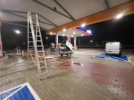 Mycie powierzchni utwardzonej na stacji benzynowej pod zadaszeniem, widoczna rozstawiona aluminiowa drabina, wąż ciśnieniowy i pojazd dostawczy. Część powierzchni umyta, widoczne zabrudzenia...