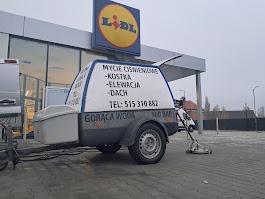 Przyczepa z napisem reklamującym usługi czyszczenia ciśnieniowego kostki, elewacji i dachów, zaparkowana na parkingu w Tarnowskich Górach przed sklepem Lidl. Widoczny wąż i urządzenie czyszczące...