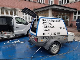 Przyczepa z napisem 'Mycie ciśnieniowe - Kostka - Elewacja - Dach' i numerem telefonu, zaparkowana na niebieskim miejscu parkingowym dla niepełnosprawnych, obok białego busa z otwartymi drzwiami...