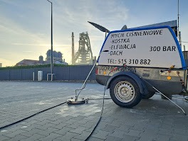 Przyczepa z urządzeniem wysokociśnieniowym do czyszczenia powierzchni na tle industrialnego krajobrazu z kominami fabrycznymi. Na przyczepie widoczny napis informujący o myciu ciśnieniowym kostki...