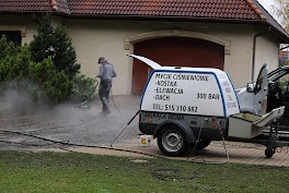 Mężczyzna w szarej bluzie czyści podjazd z kostki brukowej za pomocą myjki ciśnieniowej, obok przyczepka z reklamą usług mycia ciśnieniowego kostki, elewacji i dachów, w tle dom z czerwoną dachówką.