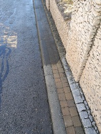 Ułożona kostka brukowa wzdłuż asfaltowej drogi, obok muru z gabionów wypełnionych jasnymi kamieniami. Widoczny brud na kostce i asfalcie.