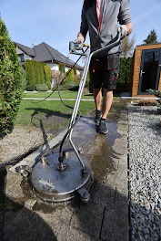 Mężczyzna w szarym swetrze i roboczych spodniach czyści kostkę brukową za pomocą profesjonalnej myjki ciśnieniowej z okrągłą dyszą, widoczny trawnik, krzewy i domek narzędziowy w tle.