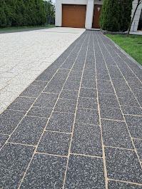 Długa, dwukolorowa ścieżka z kostki brukowej prowadząca do domu z brązową bramą garażową, z widocznym fragmentem trawnika po prawej stronie.