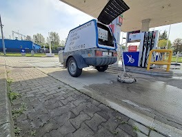 Przyczepa z urządzeniem do czyszczenia kostki brukowej na stacji benzynowej, widoczna dysza czyszcząca na kostce, błękitne logo firmy na przyczepie.