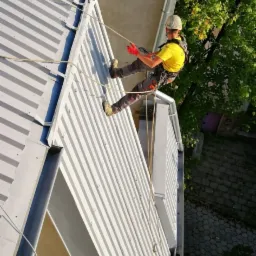 Pracownik w uprzęży, w kasku i czerwonych rękawicach, opuszcza się na linie po metalowym dachu budynku, widok z góry.