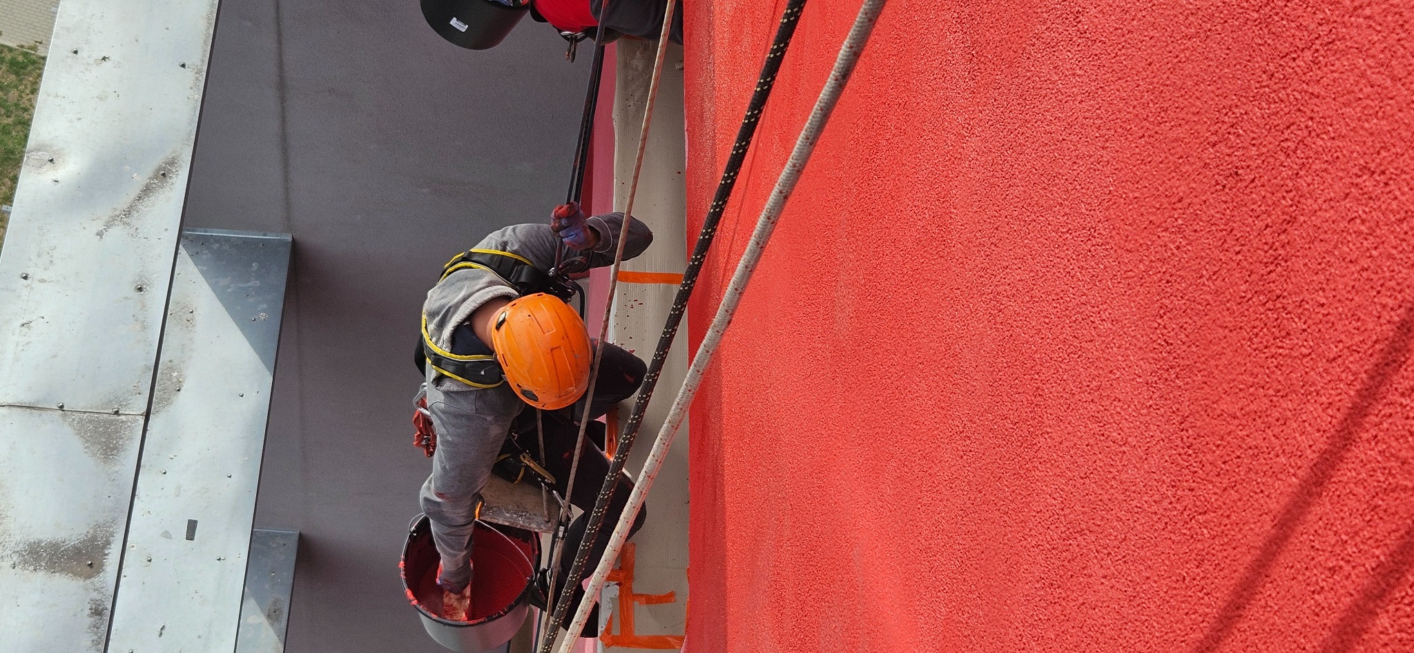 Pracownik w uprzęży, z pomarańczowym kaskiem, zawieszony na linach, maluje czerwoną elewację budynku z fakturą baranka, widok z góry.