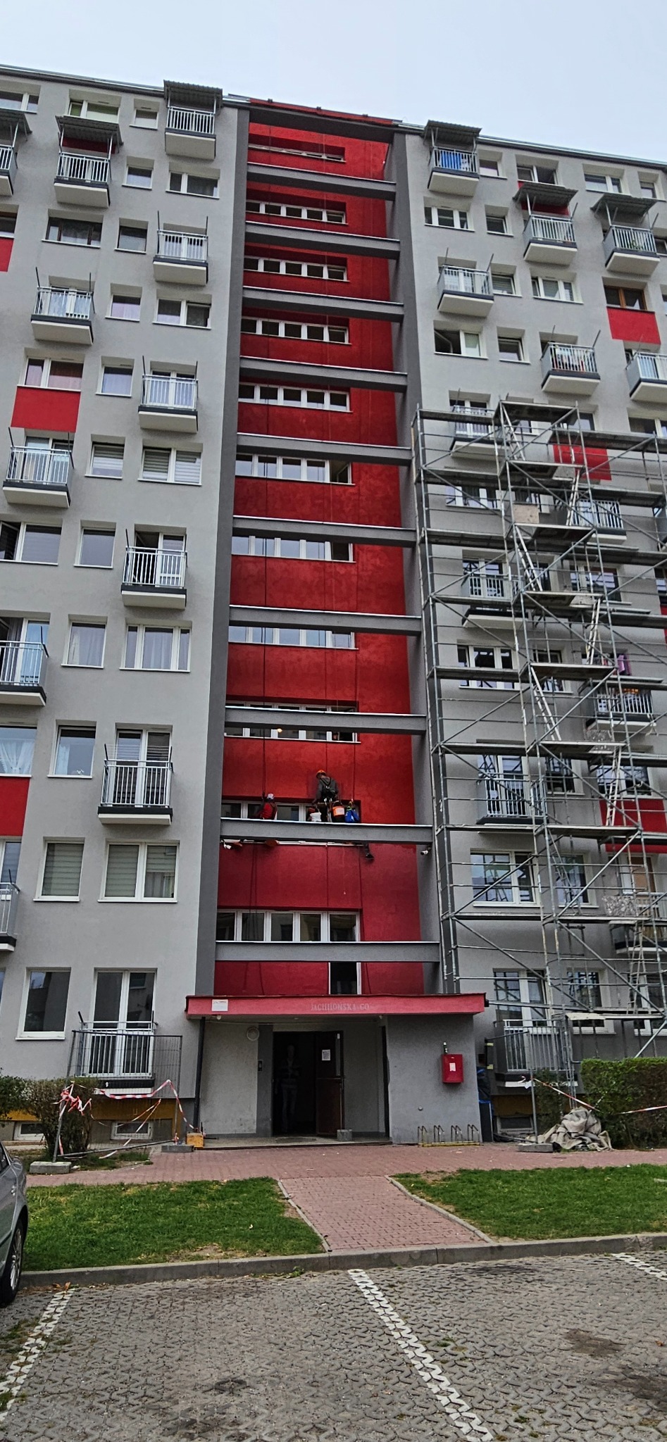 Pracownicy wysokościowi na linach malują czerwoną elewację bloku, widoczne rusztowania z prawej strony.