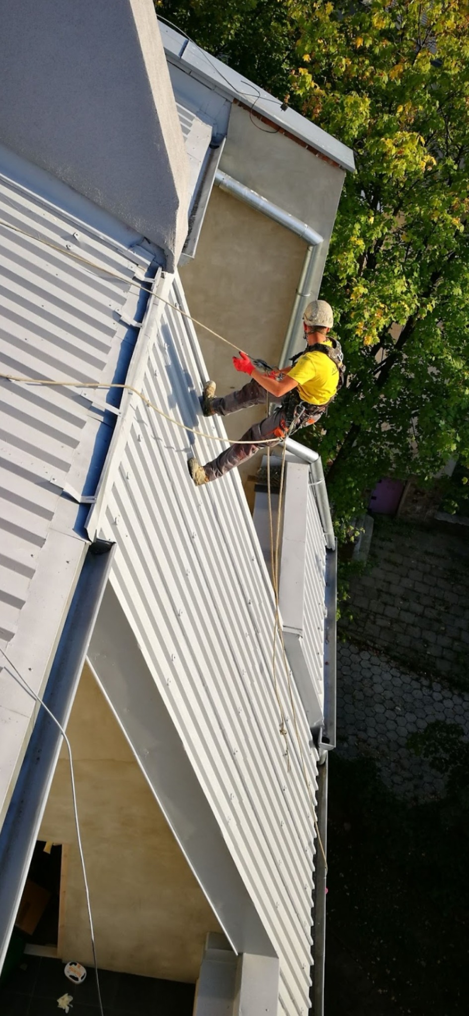 Pracownik w uprzęży, w kasku i czerwonych rękawicach, opuszcza się na linie po metalowym dachu budynku, widok z góry.