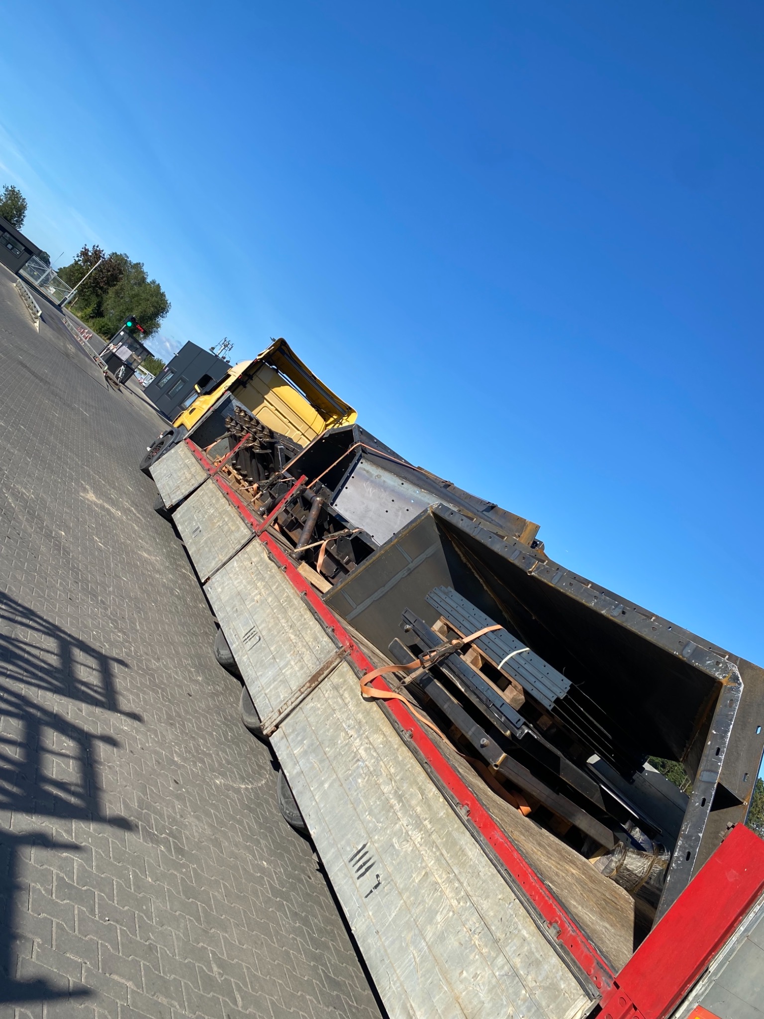 Żółta ciężarówka z otwartą skrzynią ładunkową, zabezpieczony ładunek metalowych konstrukcji i profili, błękitne niebo w tle.