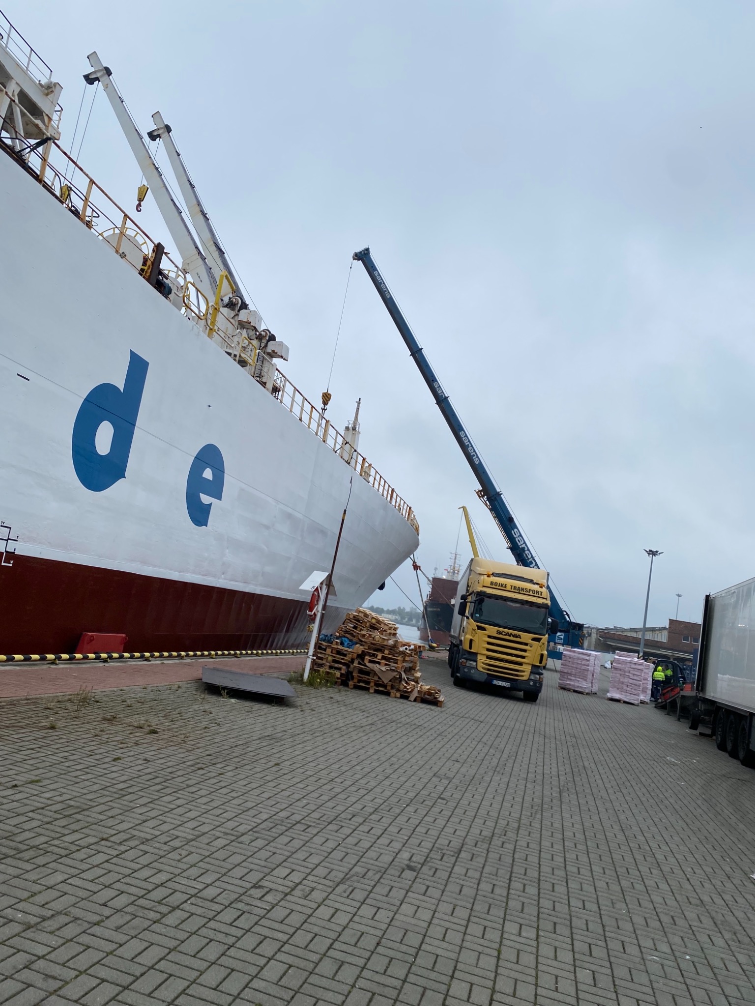 Żółta ciężarówka z dźwigiem 'Sarens' ładuje palety z towarem z dużego statku towarowego w porcie.