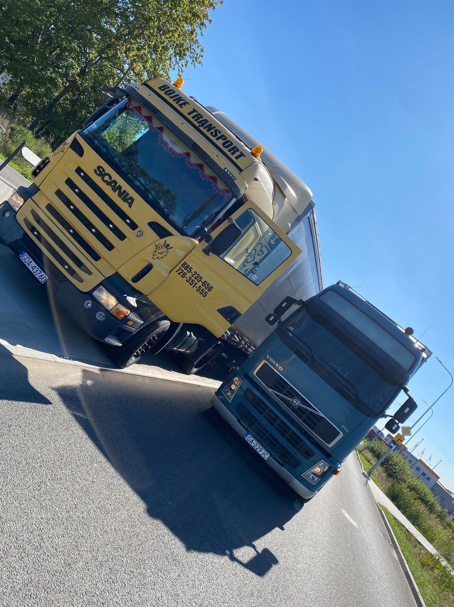 Dwa zaparkowane ciągniki siodłowe: żółta Scania z numerem telefonu i logo firmy Bojke Transport oraz niebieskie Volvo FH z numerem rejestracyjnym GDA 229JC, sfotografowane pod kątem w słoneczny dzień.