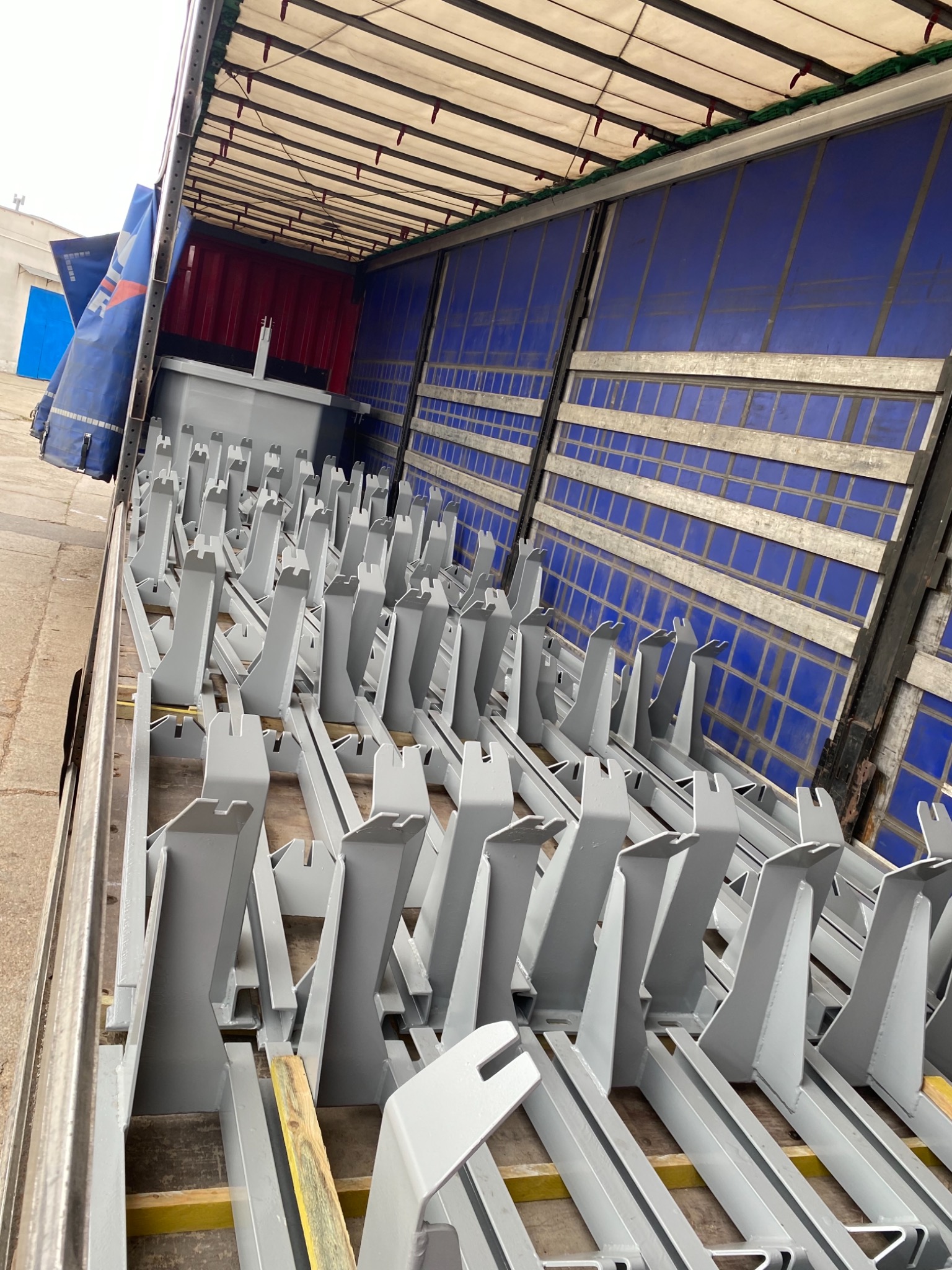 Wnętrze naczepy wypełnione szarymi, metalowymi wspornikami zabezpieczającymi ładunek, ułożonymi na żółtych belkach drewnianych, z widocznymi niebieskimi ścianami naczepy.