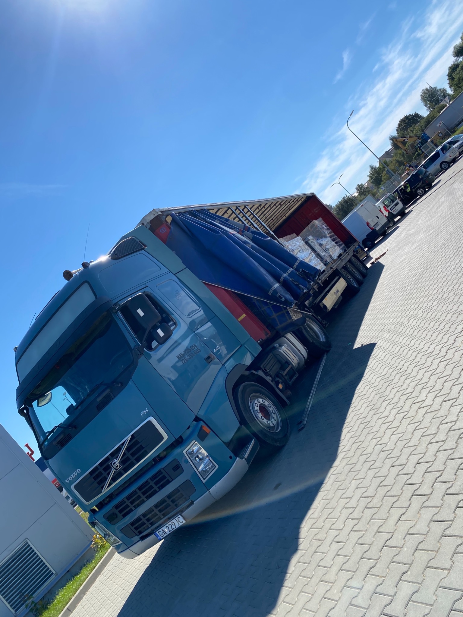 Niebieska ciężarówka Volvo FH z plandeką, wypełniona towarem, sfotografowana w Gdyni na parkingu z kostki brukowej w słoneczny dzień.