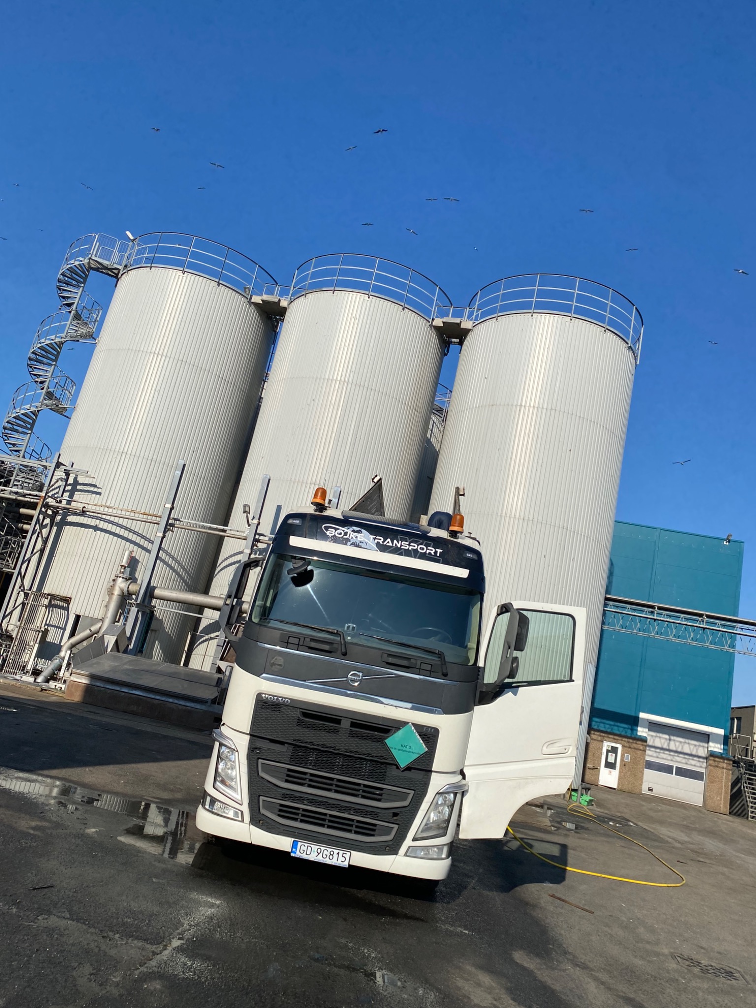 Biała ciężarówka Volvo z otwartymi drzwiami na tle trzech srebrnych silosów z spiralnymi schodami i błękitnego nieba z lecącymi ptakami.