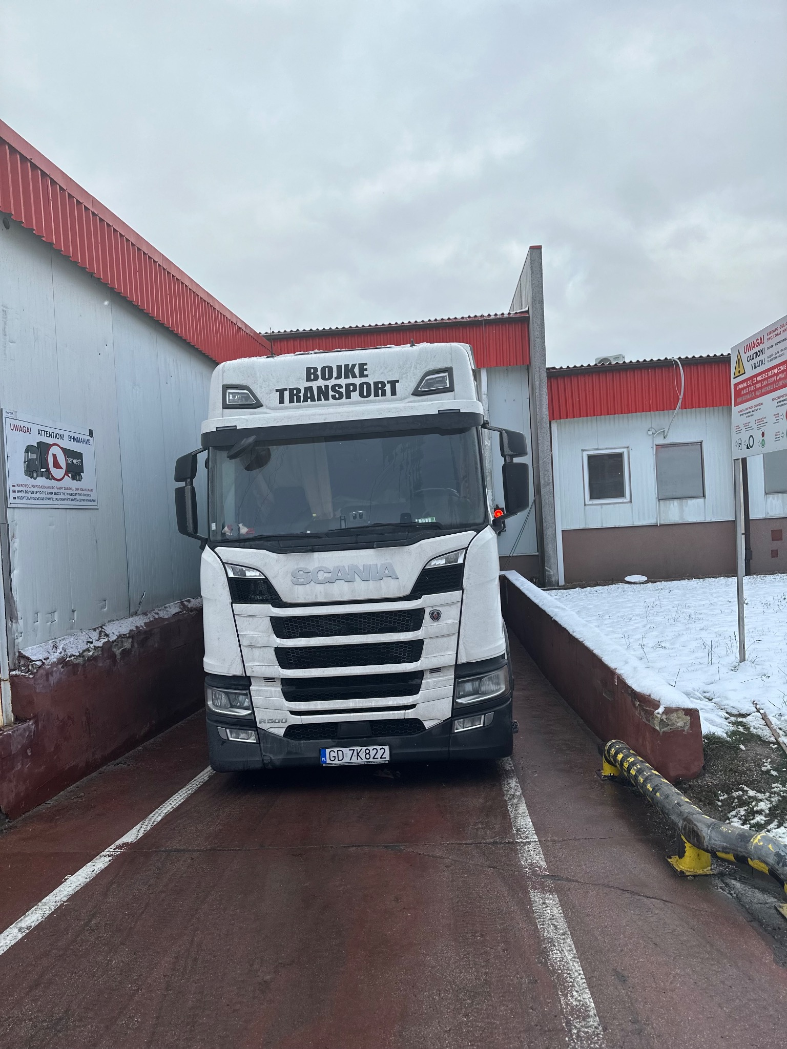 Ciężarówka marki Scania z napisem 'BOJKE TRANSPORT' na tle magazynu z czerwonym dachem i tablic ostrzegawczych.