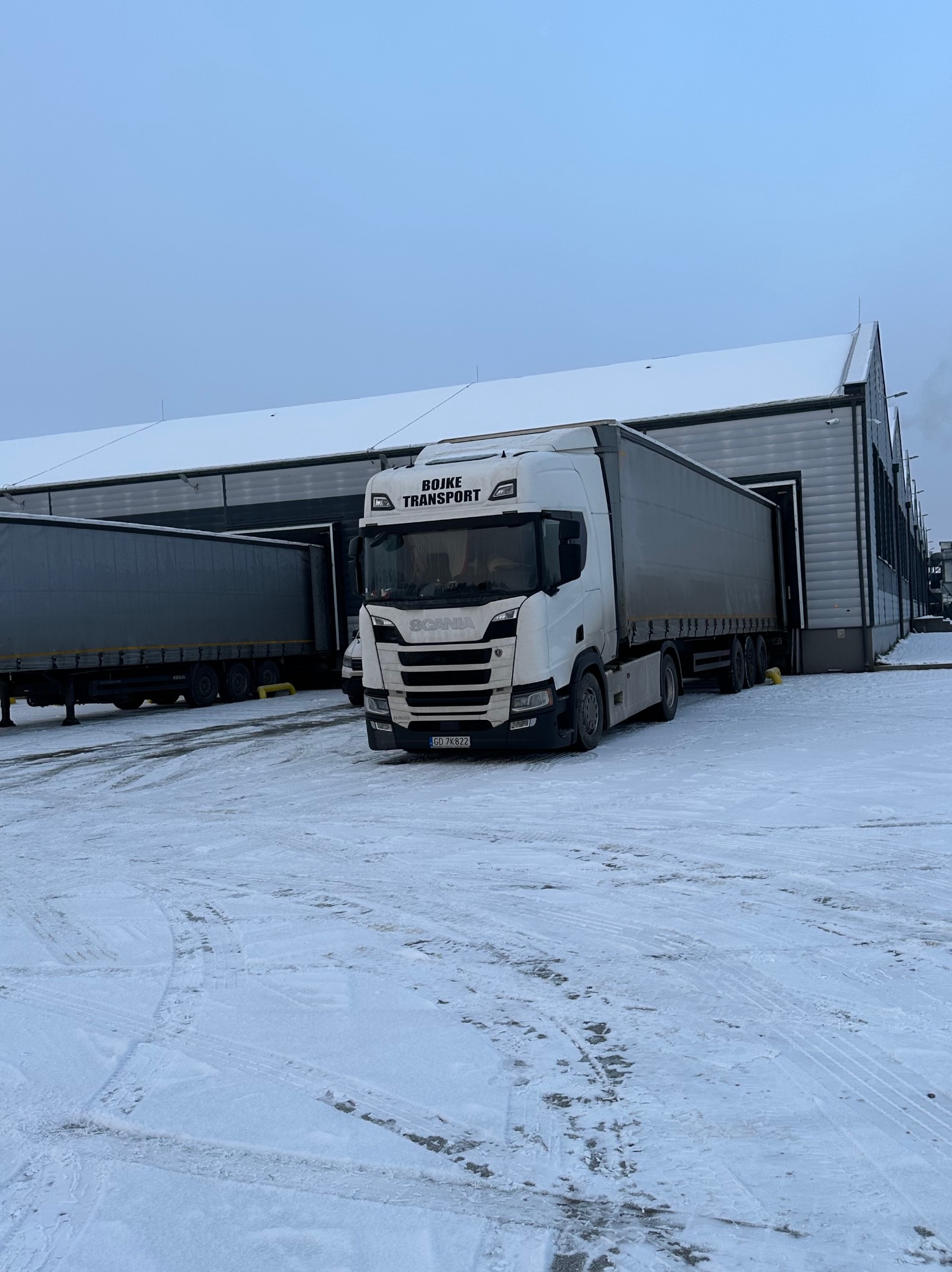 Biała ciężarówka Scania firmy Bojke Transport z naczepą wjeżdża do magazynu w zimowej scenerii, na śniegu widoczne ślady opon.