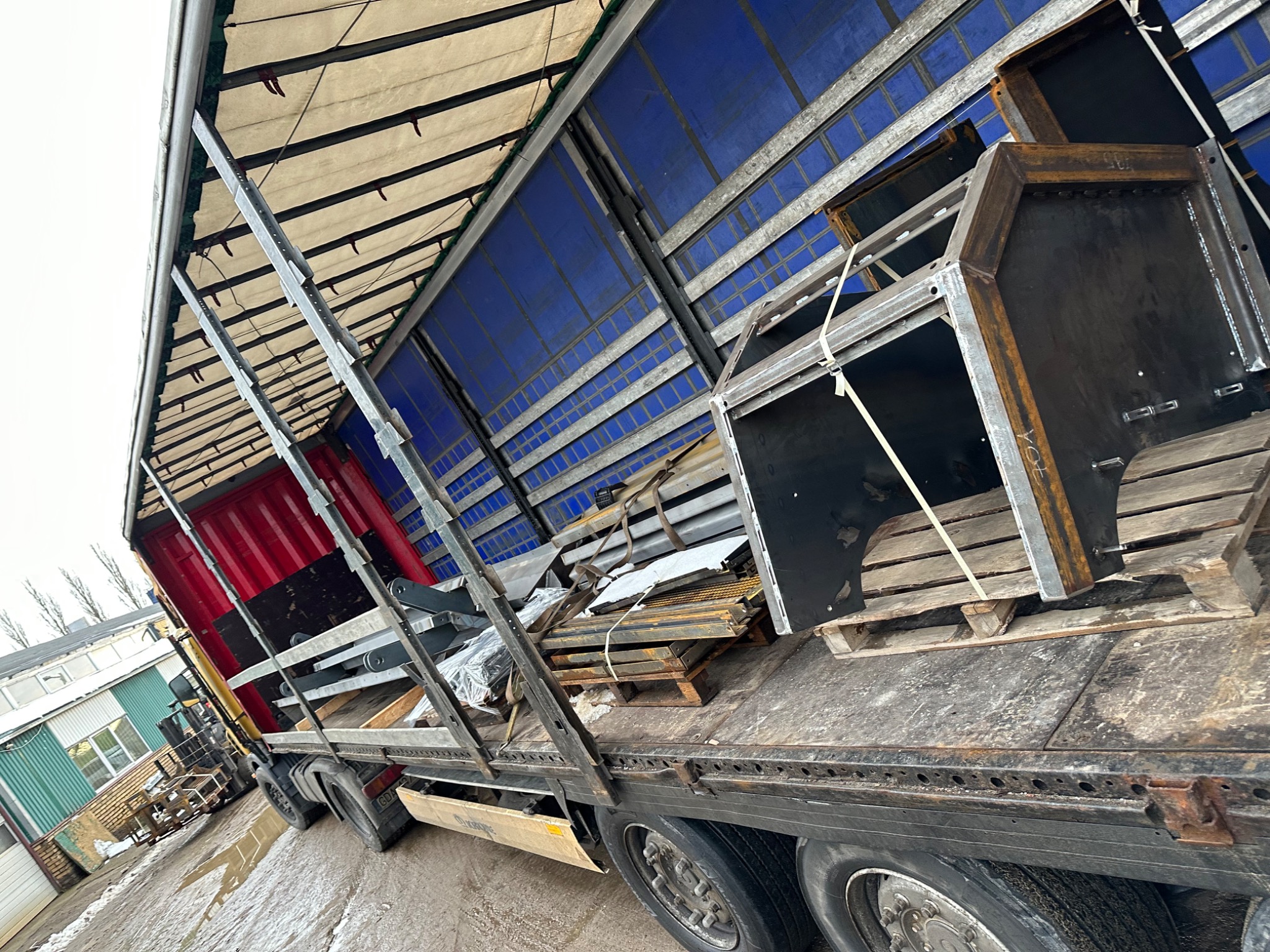 Ciężarówka z naczepą przewożąca metalowe konstrukcje i elementy na paletach, widok z boku na załadunek.