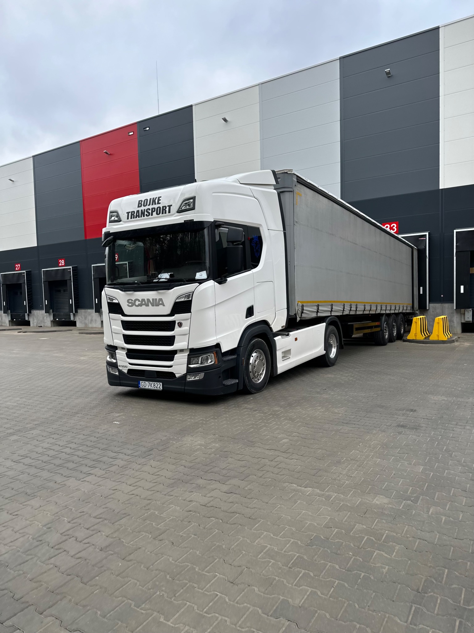 Biała ciężarówka firmy Bojke Transport z Gdyni stoi przed magazynem z rampami załadunkowymi.