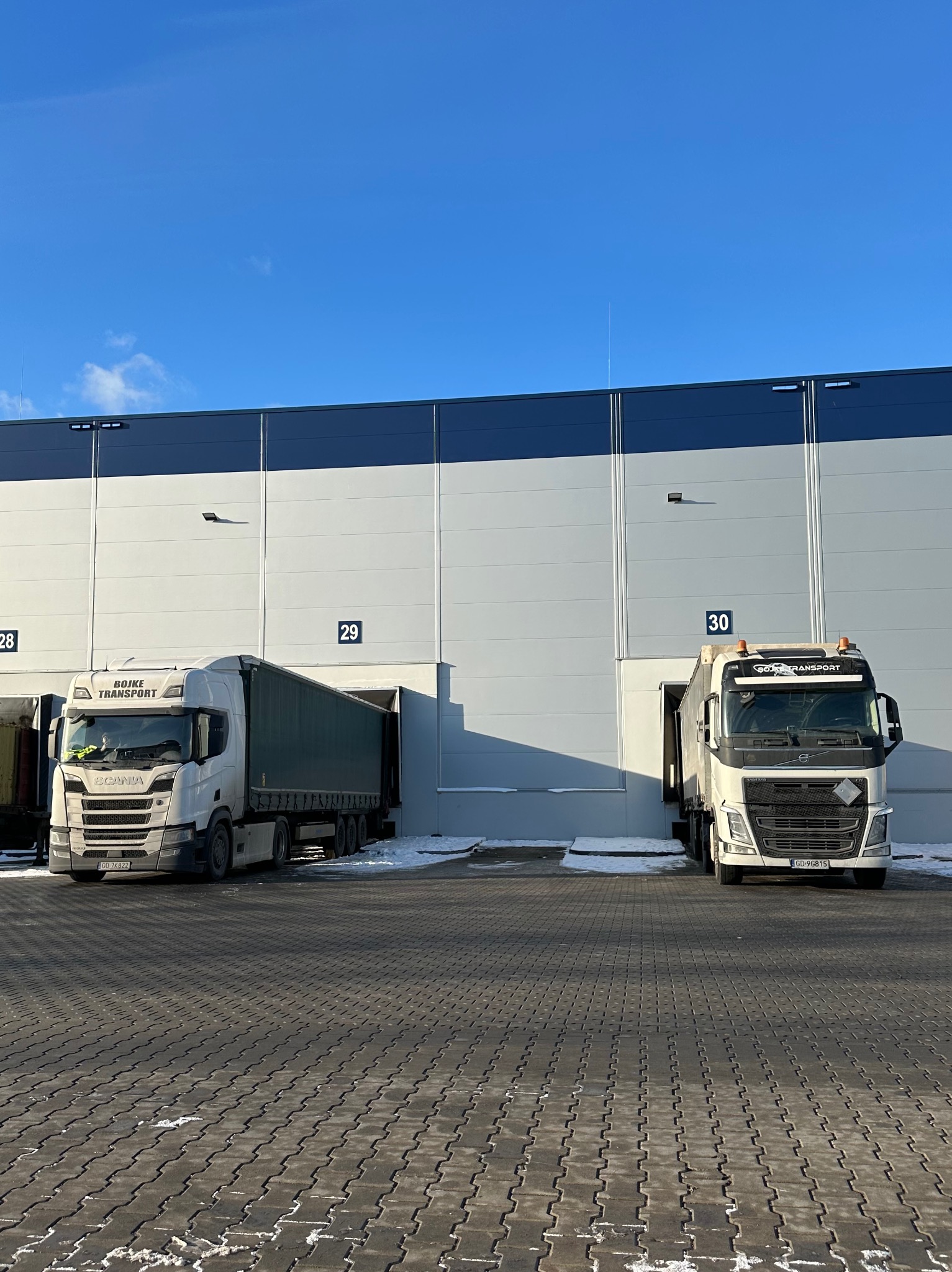 Dwa białe samochody ciężarowe marek Scania i Volvo zaparkowane przed rampami magazynowymi 29 i 30 w słoneczny dzień. Na brukowanym placu widoczne resztki śniegu.
