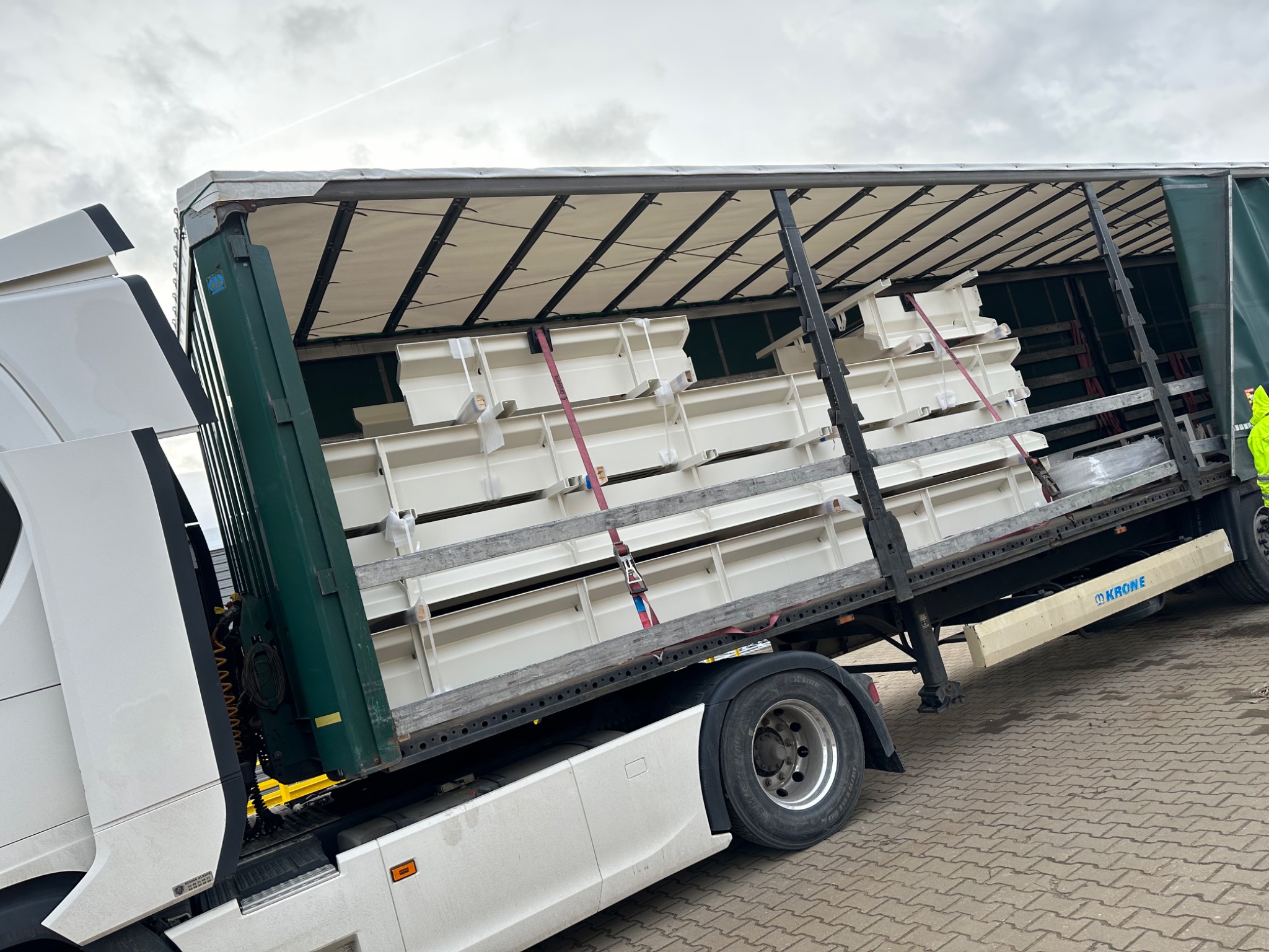 Ciężarówka z otwartą naczepą załadowana białymi, plastikowymi pojemnikami zabezpieczonymi pasami transportowymi.