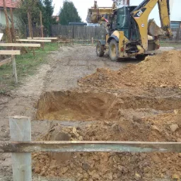 Żółta koparko-ładowarka CAT 432E wykopuje dół w ziemi, widoczne drewniane szalunki i stosy ziemi, przygotowanie terenu pod budowę.