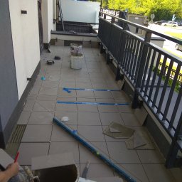 Jawor-Bud - Montaż płytek na balkonie w trakcie realizacji, widoczne narzędzia, poziomica, wiadro z zaprawą, płytki, elementy mocujące, balkon z metalową barierką, widok na zieleń za barierką.