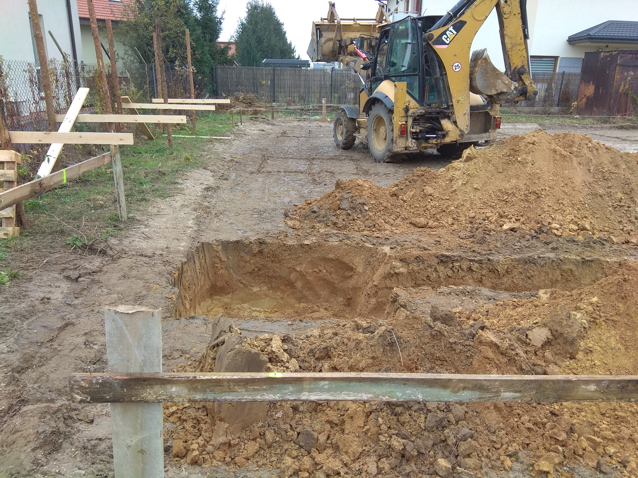 Żółta koparko-ładowarka CAT 432E wykopuje dół w ziemi, widoczne drewniane szalunki i stosy ziemi, przygotowanie terenu pod budowę.