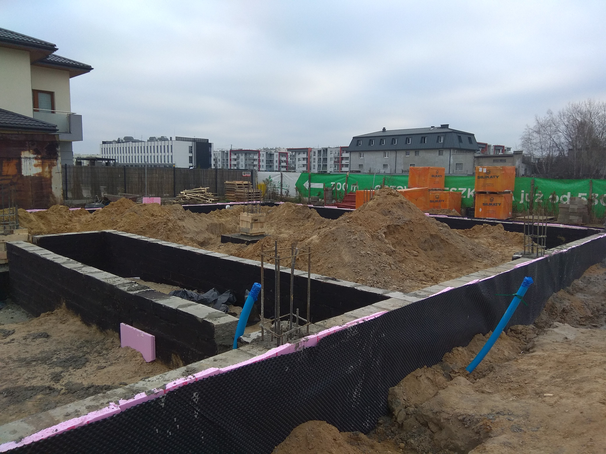 Fundamenty domu w trakcie budowy, z widoczną izolacją przeciwwilgociową, styropianem, zbrojeniami i stertami ziemi wewnątrz, na tle zabudowy miejskiej.