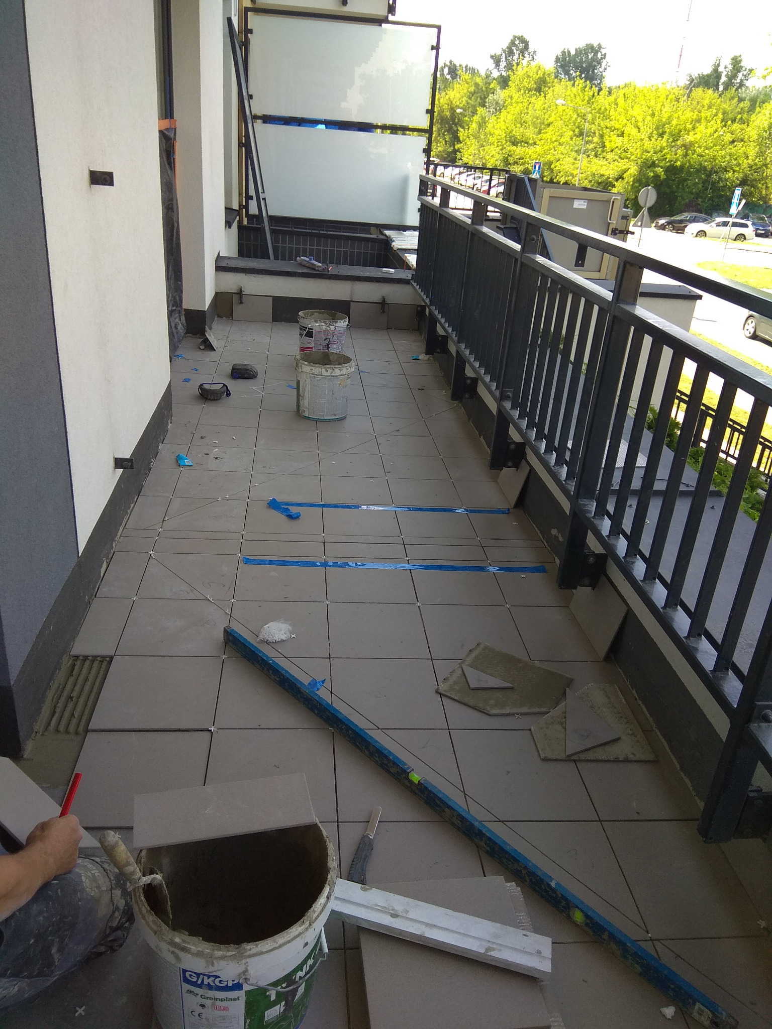 Montaż płytek na balkonie w trakcie realizacji, widoczne narzędzia, poziomica, wiadro z zaprawą, płytki, elementy mocujące, balkon z metalową barierką, widok na zieleń za barierką.