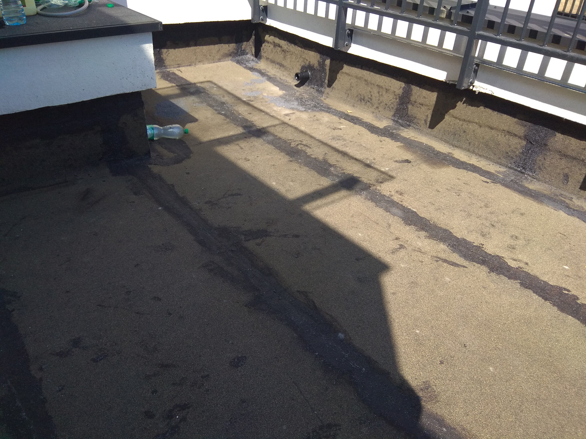 Widok z góry na płaski dach pokryty papą z widocznymi łączeniami, fragmentem metalowej balustrady i plastikową butelką w rogu.