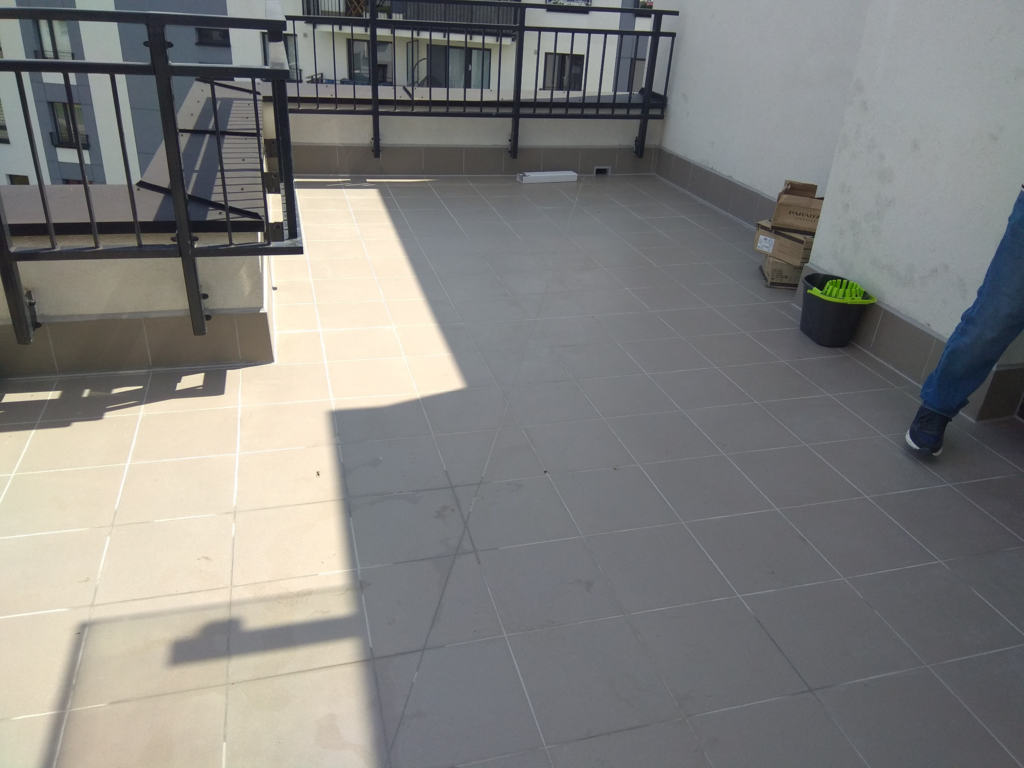 Wyłożony szarymi płytkami taras z czarną metalową balustradą, widoczne kartony i wiadro z mopem przy ścianie, fragment nogi osoby w jeansach w prawym dolnym rogu.