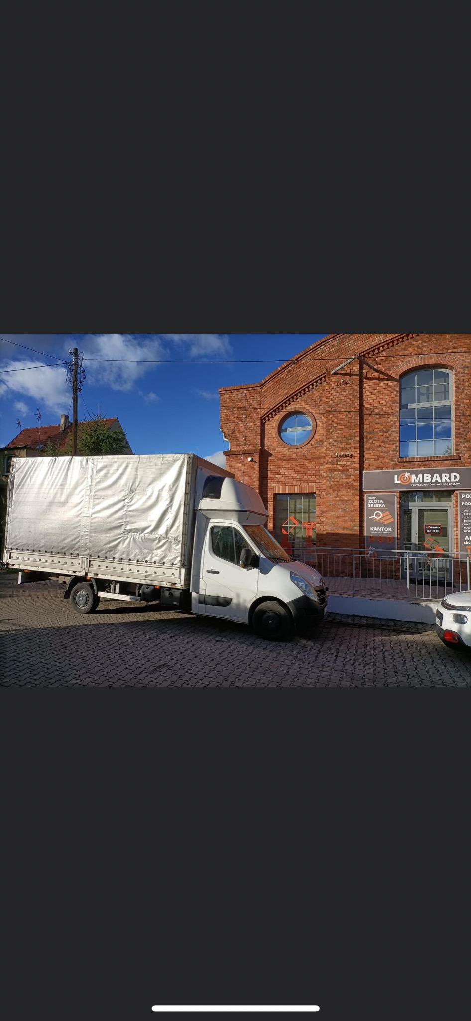 Biały samochód dostawczy z plandeką zaparkowany na kostce brukowej przed budynkiem z czerwonej cegły z szyldem 'LOMBARD'.