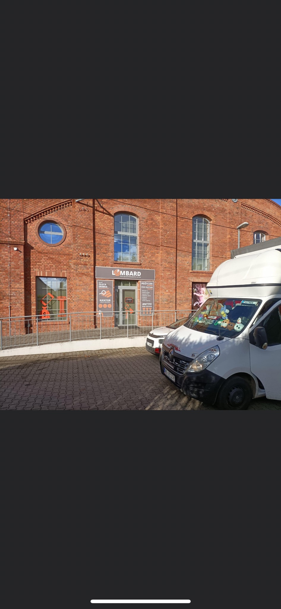 Biały bus Renault Master zaparkowany przed budynkiem lombardu z cegły, ozdobiony naklejkami i pluszakami na przedniej szybie.