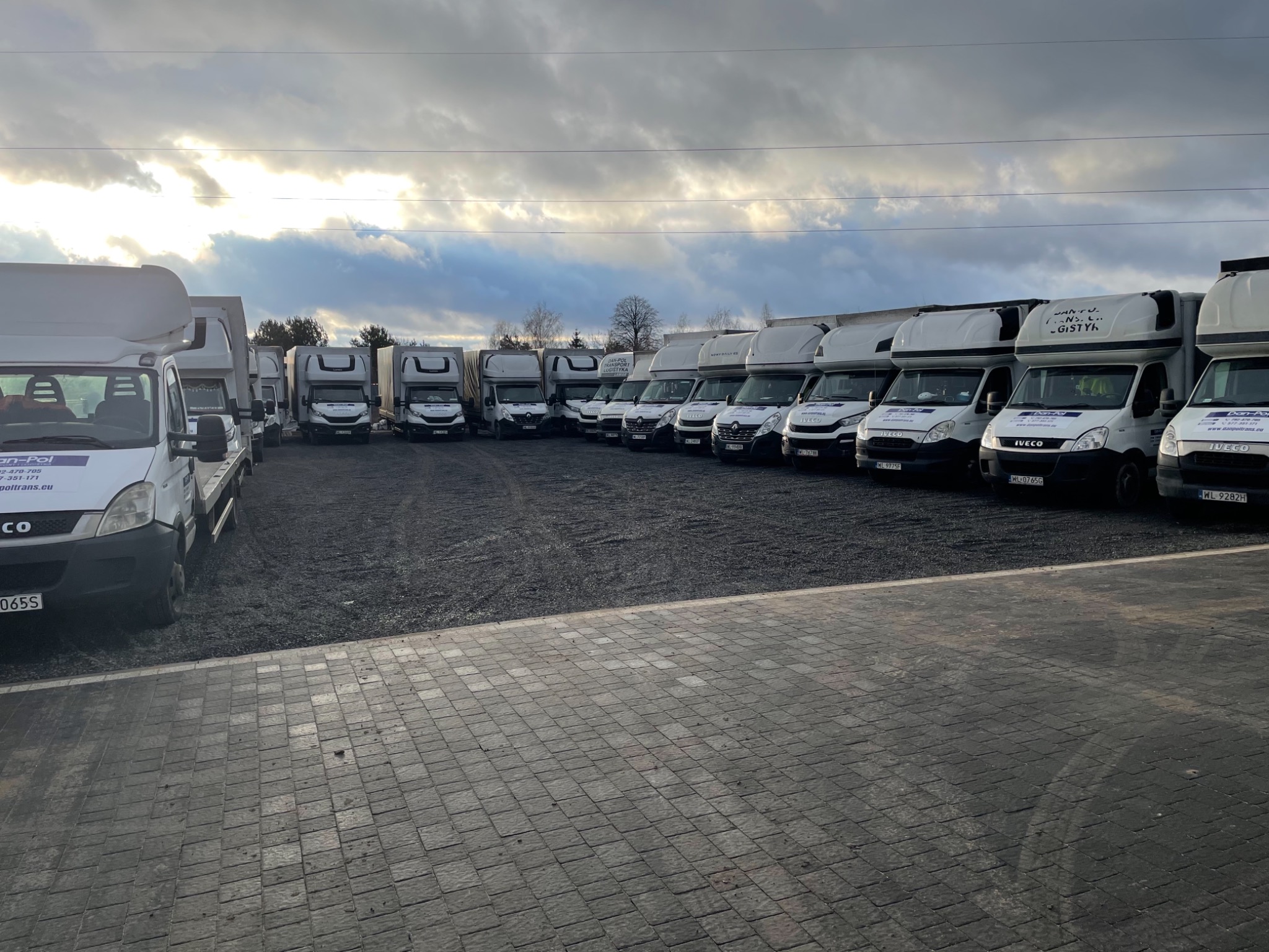 Flota białych samochodów dostawczych Iveco z zabudową kontenerową, zaparkowanych rzędem na żwirowym placu, z widocznymi oznaczeniami firmowymi Dan-Pol Transport Logistyka.