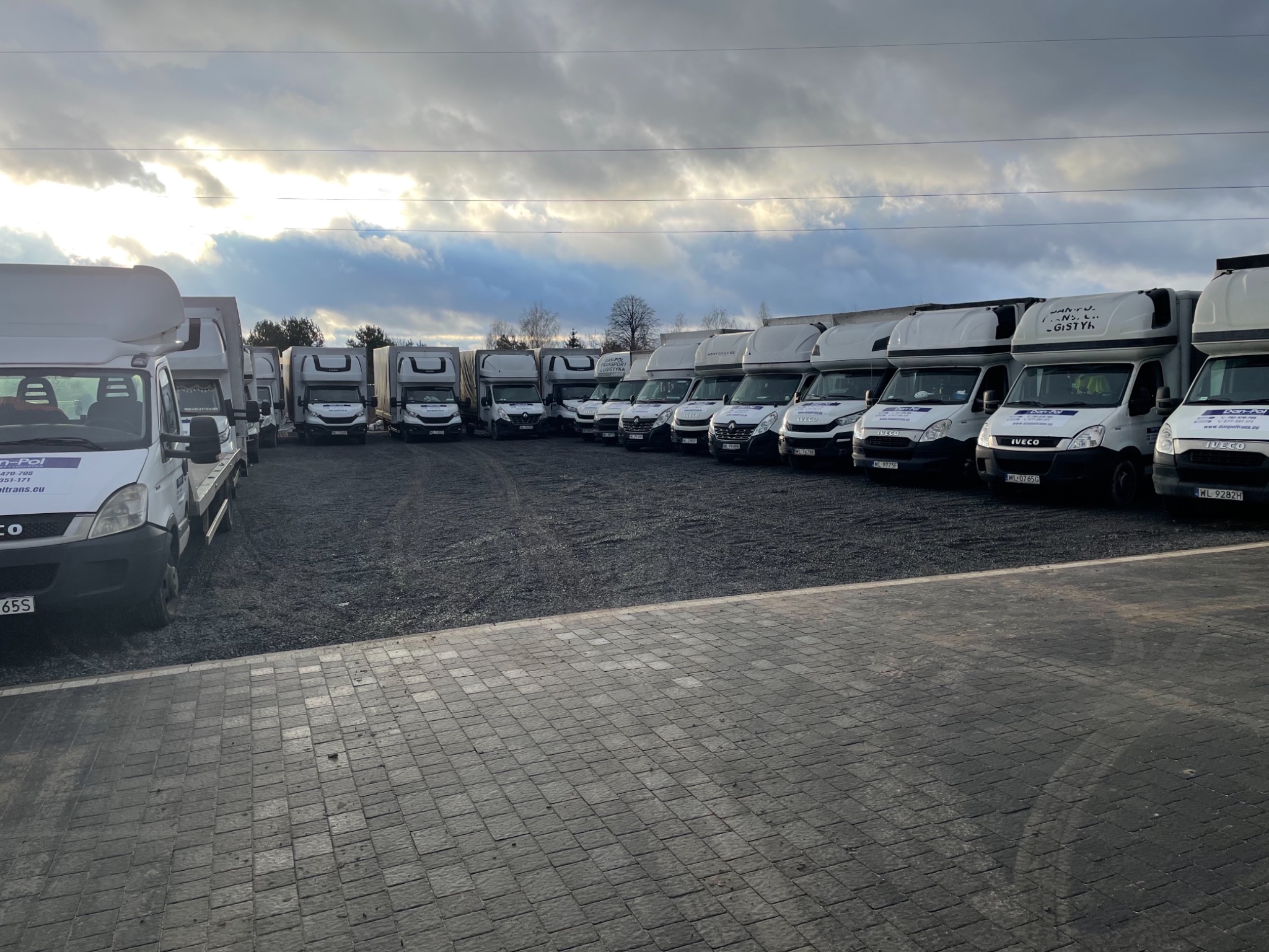 Flota białych samochodów dostawczych Iveco firmy transportowej Dan-Pol zaparkowana na żwirowym placu, pochmurne niebo w tle.