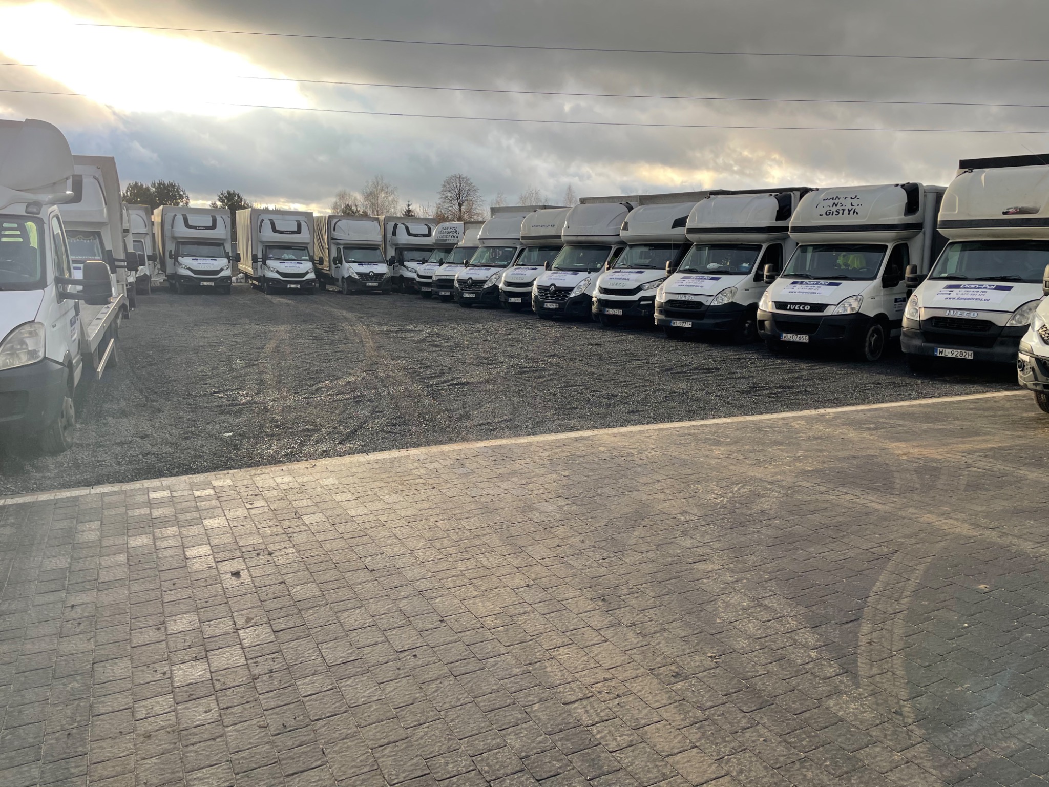 Flota białych samochodów dostawczych z nadbudową, ustawionych w rzędzie na żwirowym placu, z logo firmy transportowej na każdym pojeździe, sfotografowana w pochmurny dzień.